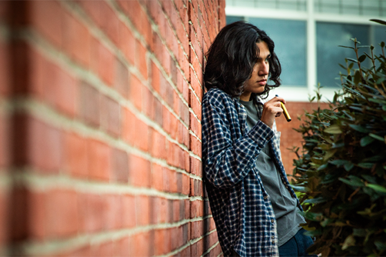 Un adolescente con el pelo largo apoyado contra una pared de ladrillos, sosteniendo una herramienta de vapeo. Esta publicación de blog describe los riesgos de vapear para los adolescentes.